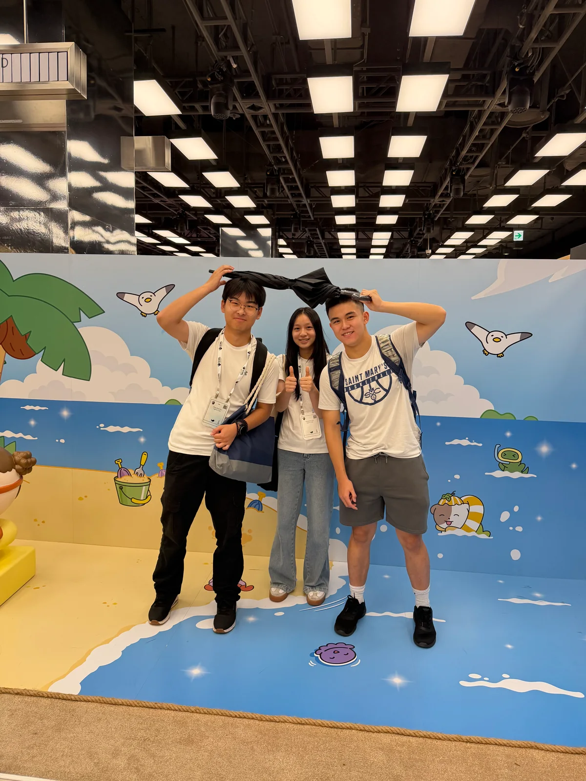 Three students smiling together beneath an umbrella in front of a colorful conference display.